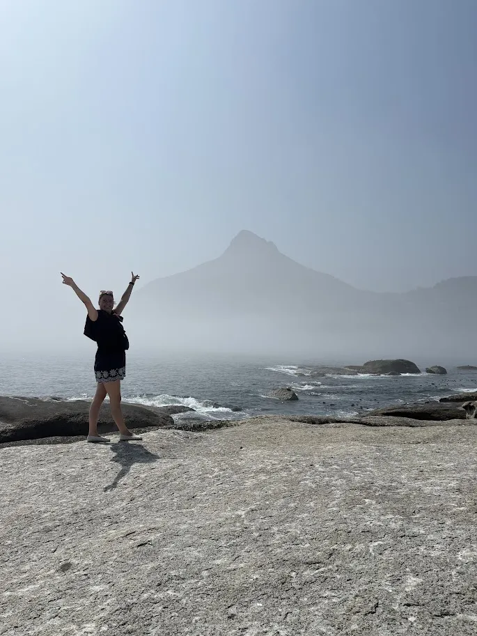 Een vrouw met haar armen omhoog op de rotsen aan zee, met Lion's Head in de mist